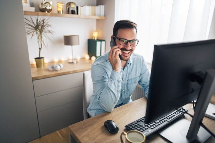 A Young Caucasian Man in Glasses Sits at a Desktop Computer in His Home Office While Talking on his Cell Phone and Smiling