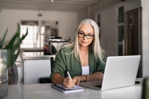 A woman sitting at a table and writing on a notepad with a laptop in front of her.